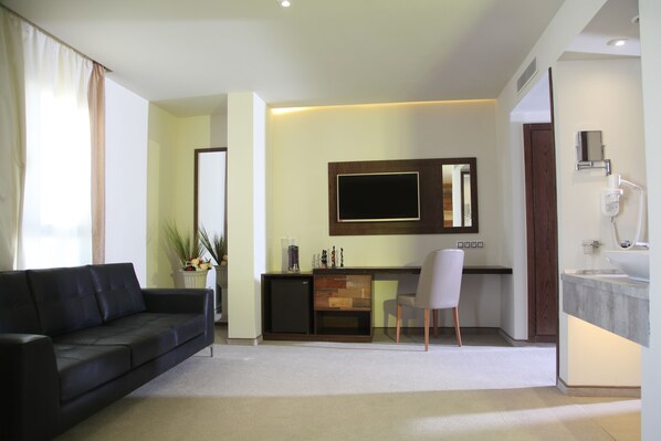 A modern living room with a black sofa, a small desk, a TV, and a bathroom with a sink and mirror. - Business Hotel Sfax (Sfax)