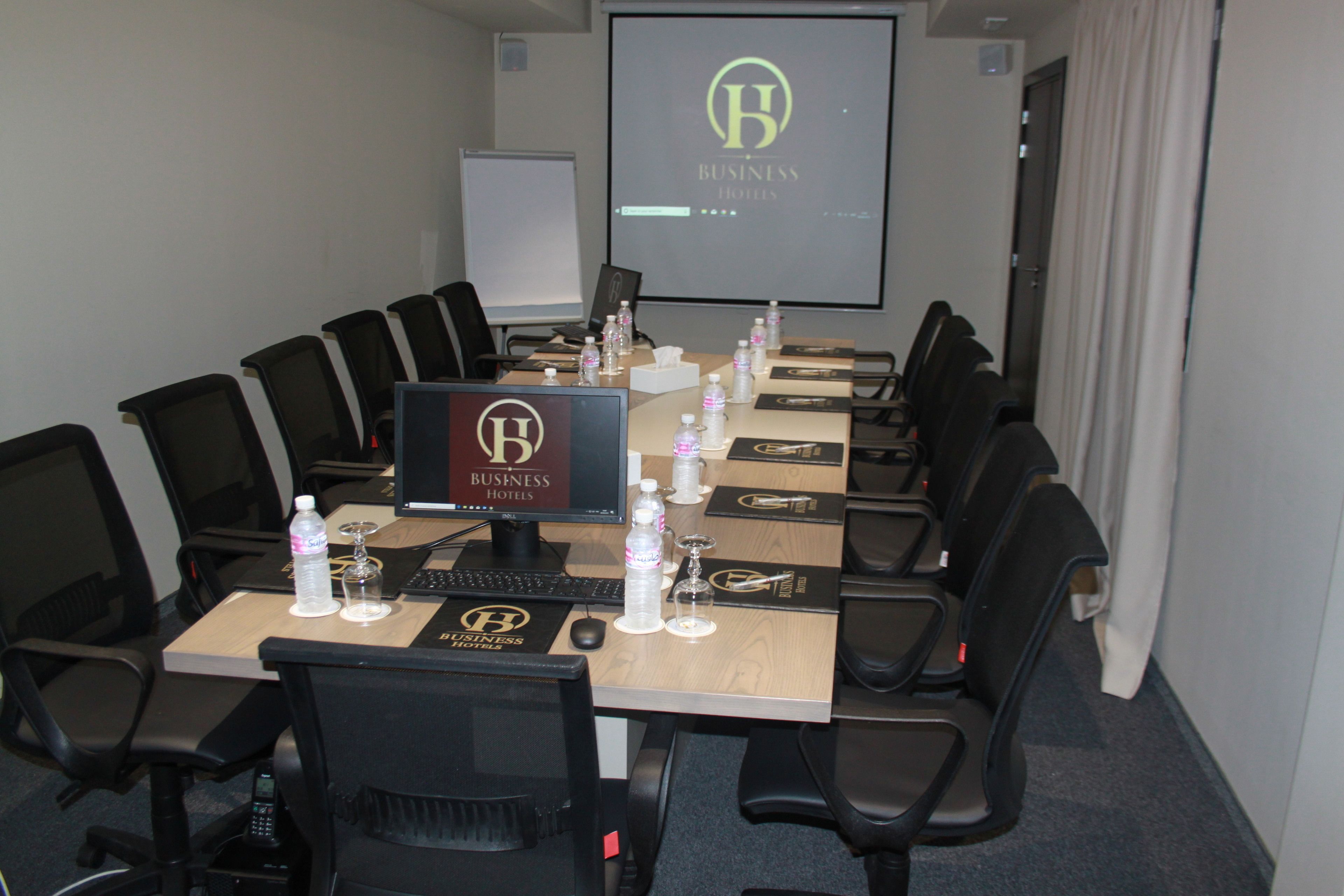 Un salón de conferencias con una larga mesa, sillas negras, una pantalla de proyector y un monitor que muestra el logotipo de Business Hotels.