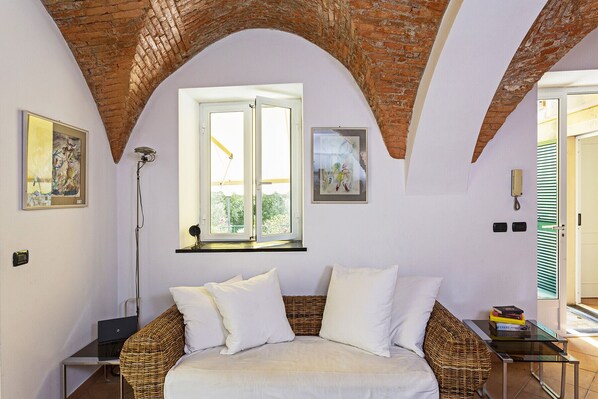 A room with a bed, a wicker chair, a small table, and a window with a view of greenery.