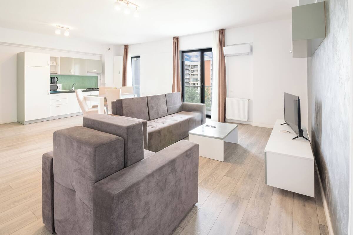 Una sala de estar moderna con un sofá seccional gris, una mesa de centro blanca y un televisor de pantalla plana sobre un mueble blanco. Se ve un área de cocina con armarios blancos y un salpicadero verde a través de una puerta abierta.