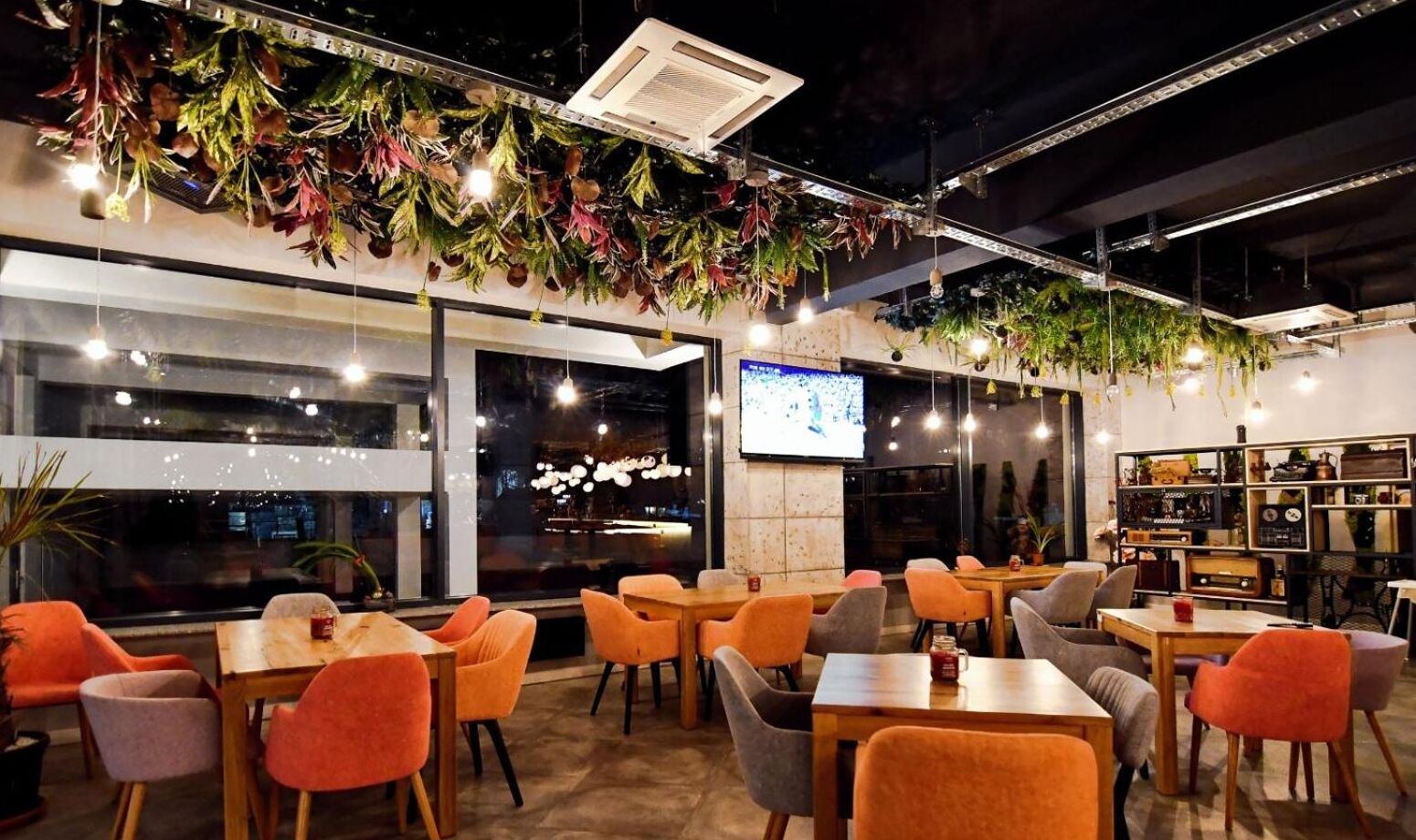 A modern indoor dining area with orange and gray chairs, a large TV screen, and hanging plants.