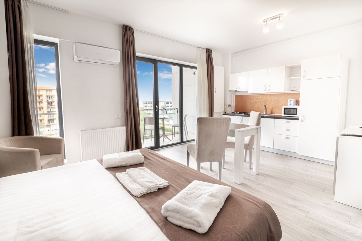 A hotel room with a bed, a view of the city, a kitchenette, and a balcony.