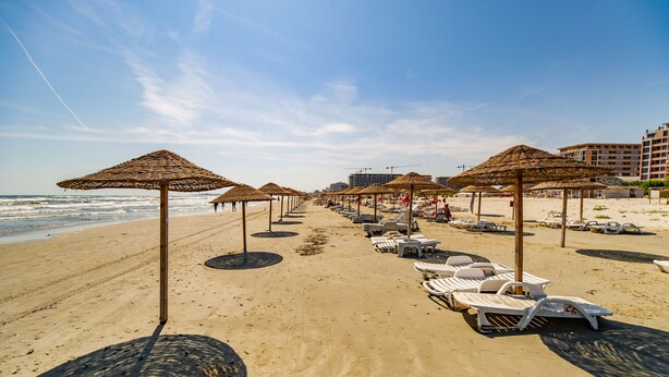 A beach with rows of sun loungers and thatched umbrellas, a clear sky, and a building in the background. - Miramare Residence (Navodari)