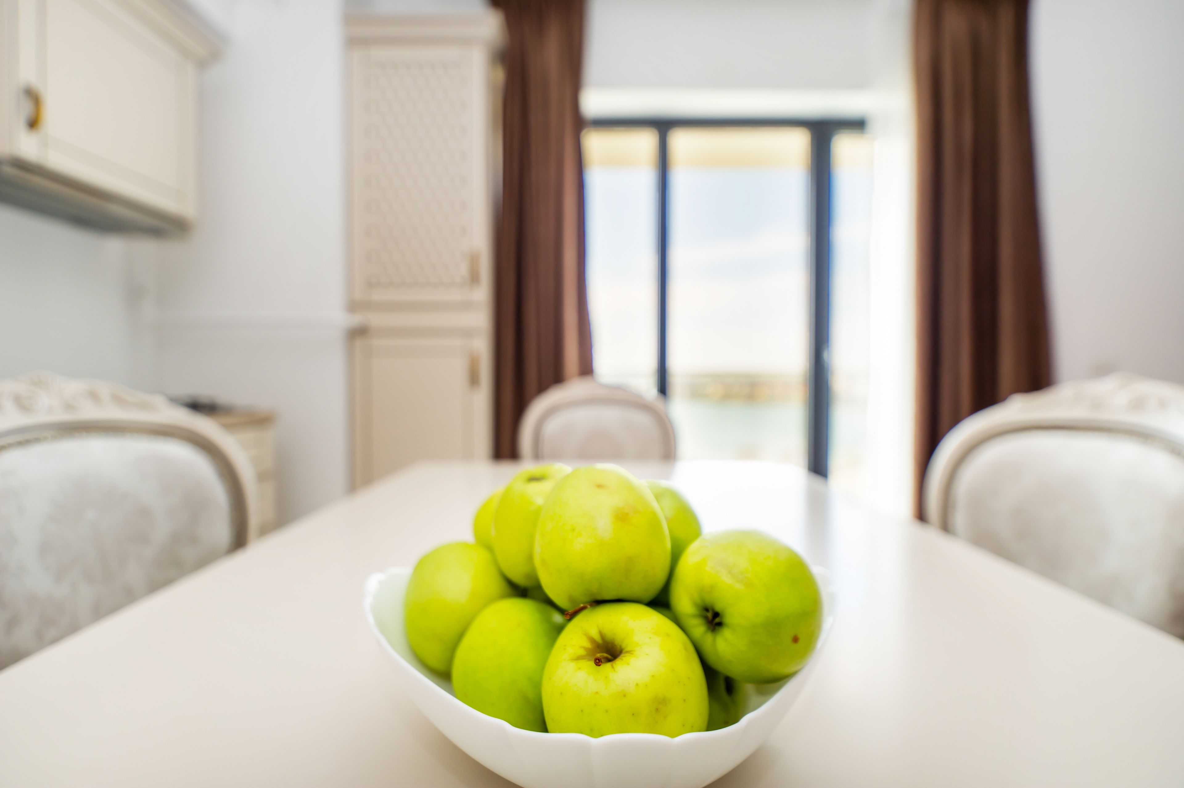 A white dining table with a bowl of green apples in the center.