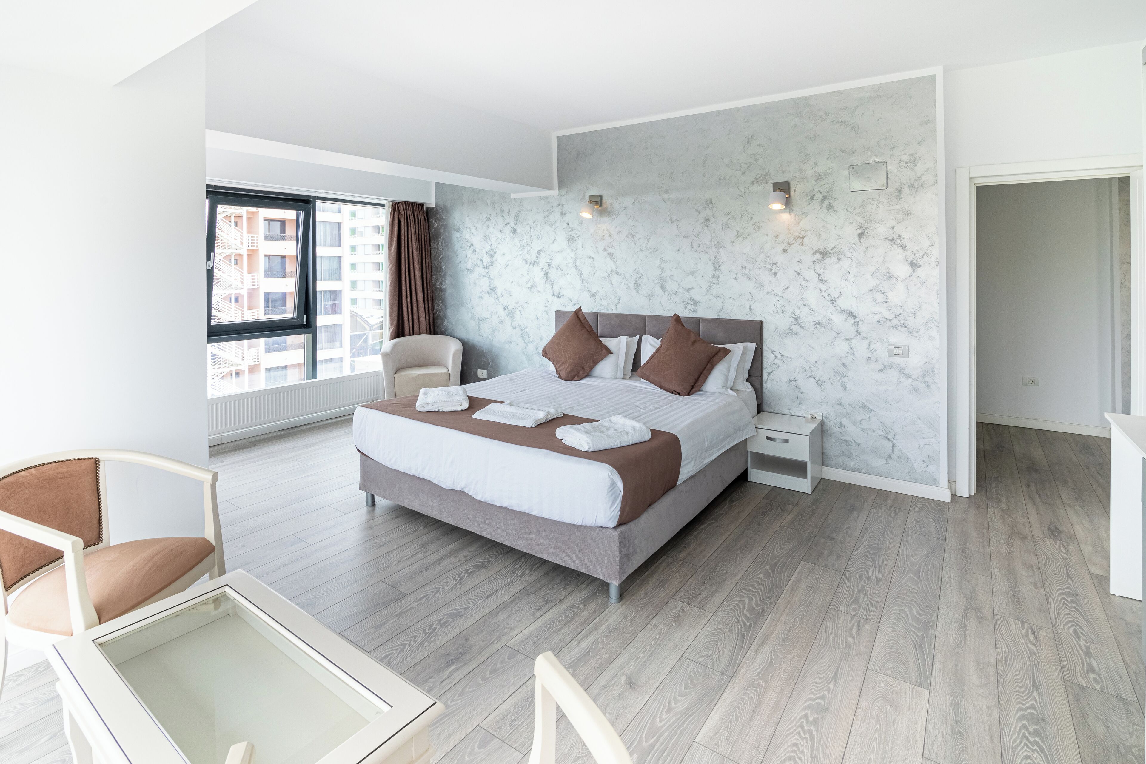 A modern hotel room with a large bed, a sitting area with a chair, a small table, and a view of the city through a window.