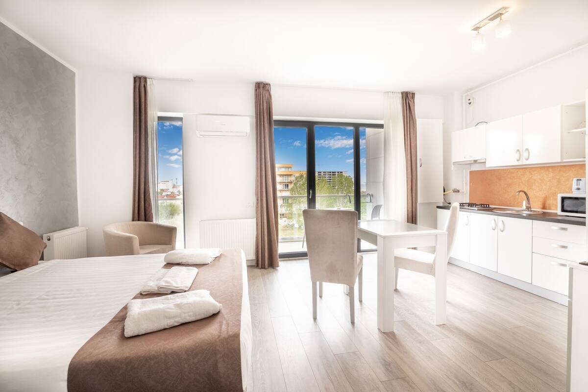 Una habitación de hotel moderna con cama, mesa de comedor, zona de cocina y vistas a la ciudad.