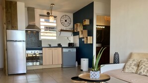 A modern kitchen with a refrigerator, stove, and dishwasher. There is a dining area with a table and chairs, and a living room with a sofa and a plant. - The lagoon terrace (Saint Leu)