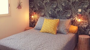 A bedroom with a bed, a wall-mounted plant, a wooden stool, and a window. - The lagoon terrace (Saint Leu)