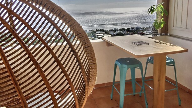 A balcony with a view of the sea, a round wicker chair, a table, and two blue stools. - The lagoon terrace (Saint Leu)