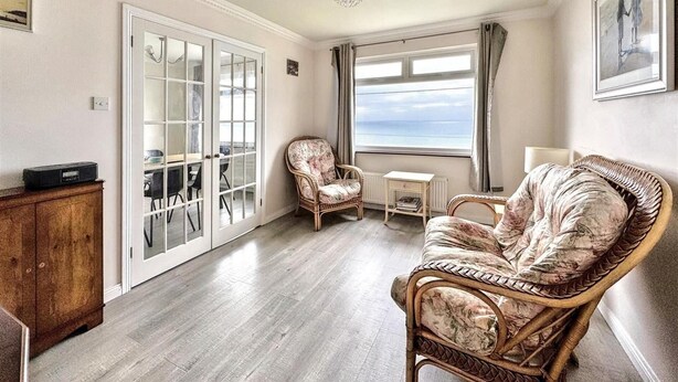 Een kamer met een houten vloer, een rieten stoel, een bank, een tafeltje, een kast en uitzicht op zee door een raam.