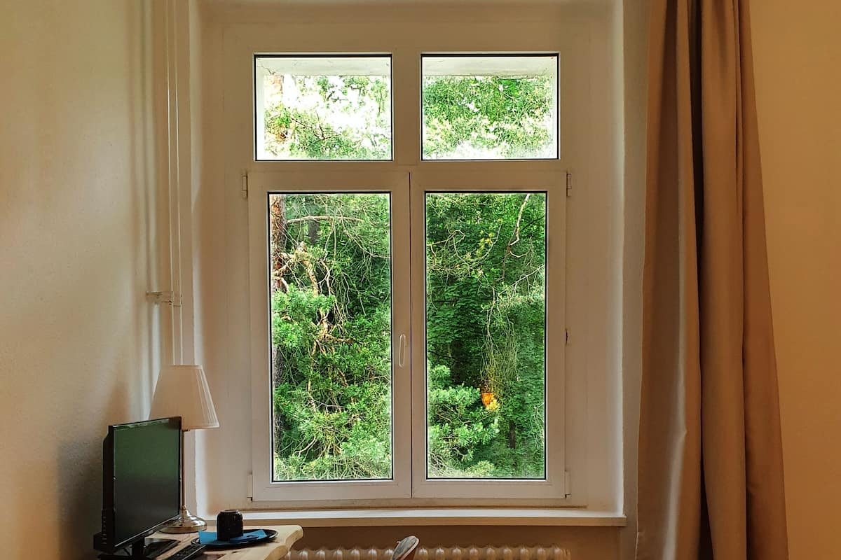 A bedroom with a bed, a desk with a computer, a window with curtains, and a view of greenery outside.