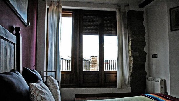 A bedroom with a bed, a window with curtains, a view of a balcony, and a stone wall. - Rural house for 4 people (Ayerbe)