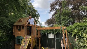 A wooden playhouse with a slide and climbing wall, surrounded by a garden with a stone border and small bushes. - The Deeping Stage (Peterborough)