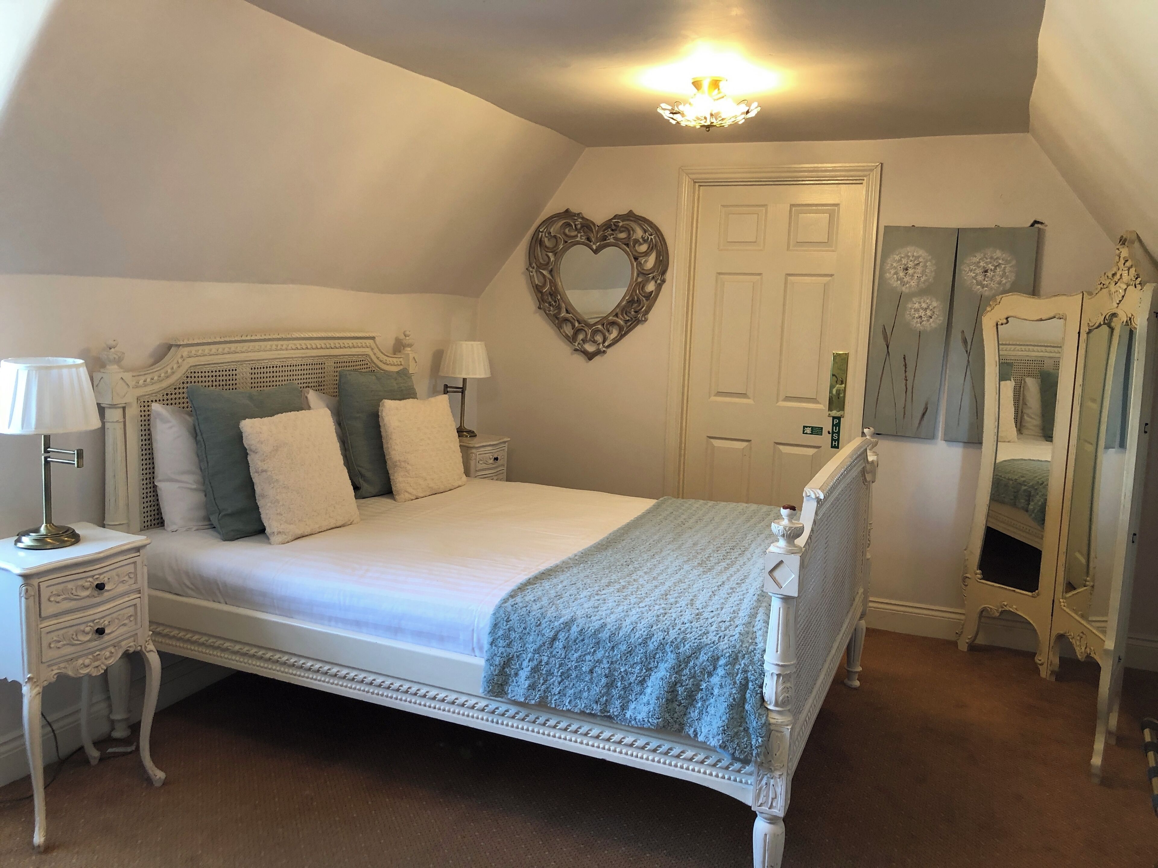 Un dormitorio con cama con dosel, una mesita de noche con lámpara, cómoda, espejo y puerta decorada con un adorno en forma de corazón.