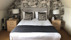 A bedroom with a sloped ceiling, a bed with striped bedding, two zebra-patterned pillows, two bedside lamps, a nightstand with a lamp, a radiator, and a telephone. - The Deeping Stage (Peterborough)