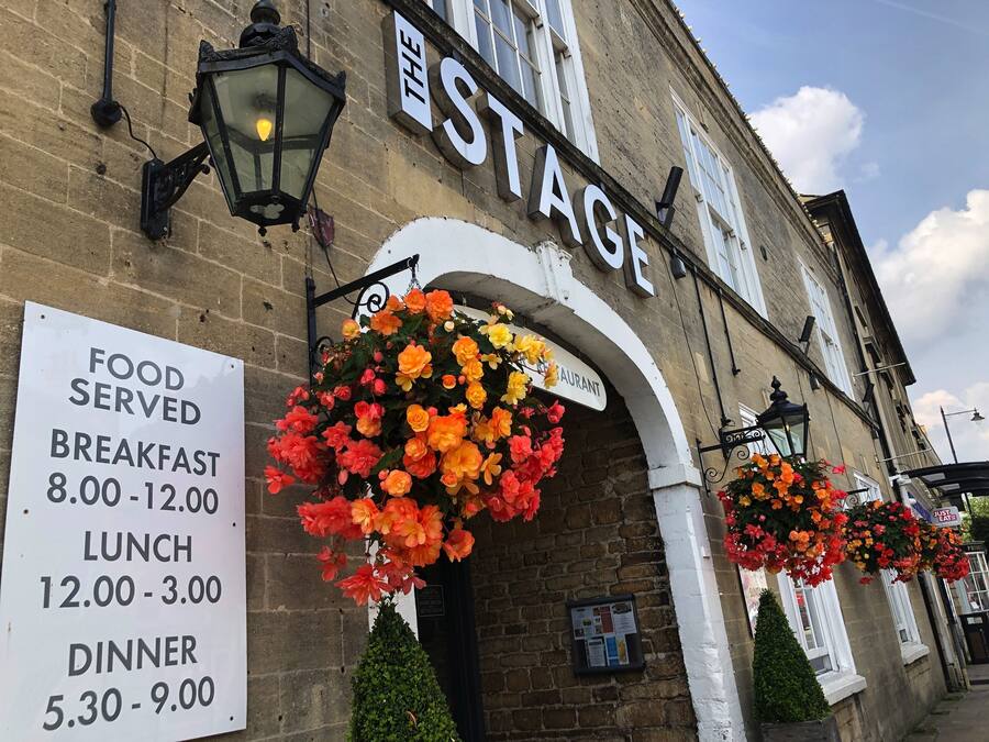 Un edificio con un cartel que dice THE STAGE y un panel de menú que anuncia horarios de desayuno, almuerzo y cena.