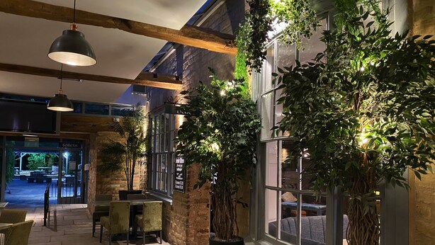 An indoor area with stone walls, wooden beams, and potted plants. There are tables and chairs, and a sign indicating a seating area. - The Deeping Stage (Peterborough)
