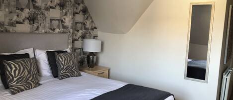Une chambre à coucher avec un lit, des oreillers, une table de chevet avec une lampe, un miroir et un mur orné d’un motif floral.