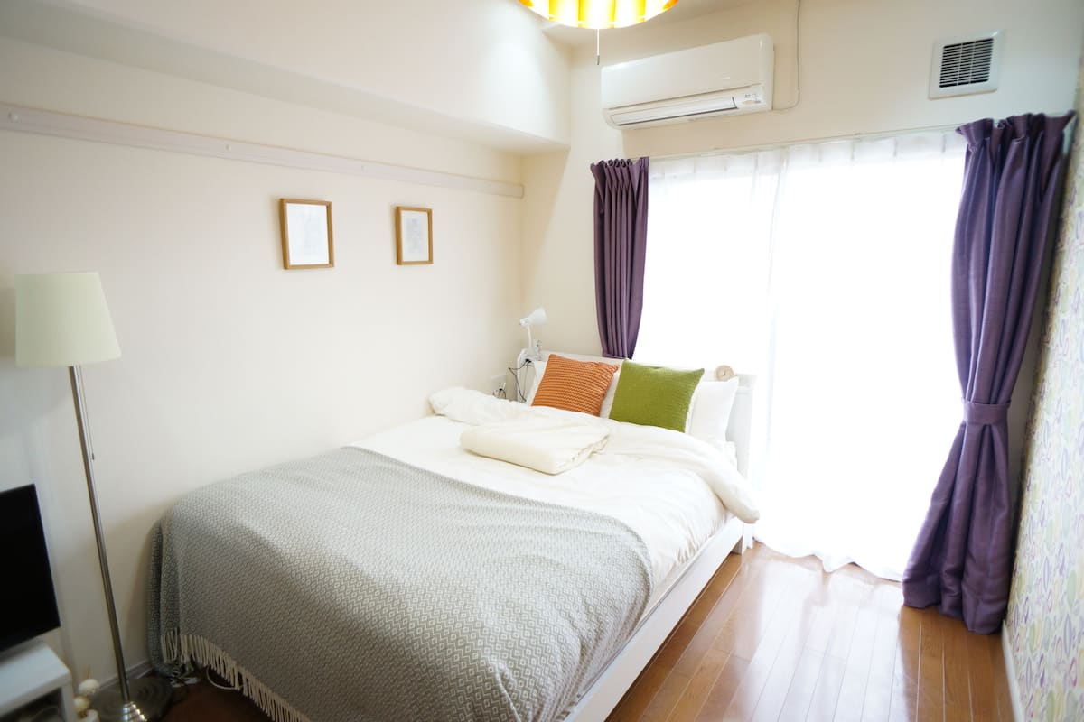 A neatly made bed with pillows and a blanket, a bedside lamp, and a flat-screen TV on a stand.