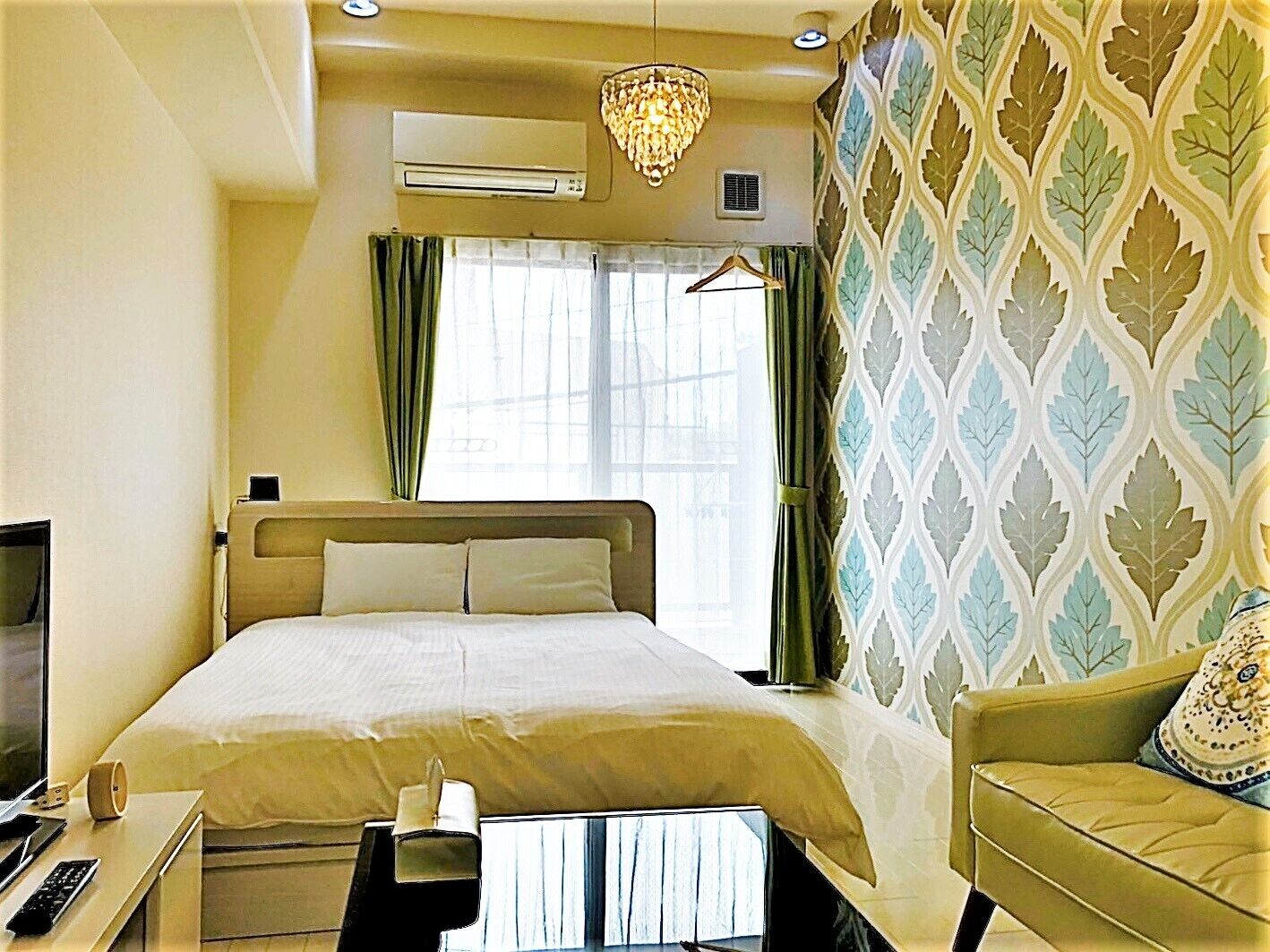 A hotel room with a bed, a desk with a chair, a TV, a chandelier, and a patterned wall.