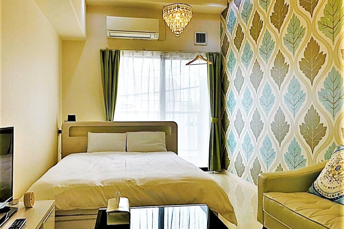 A hotel room with a bed, a desk with a chair, a TV, a chandelier, and a patterned wall.