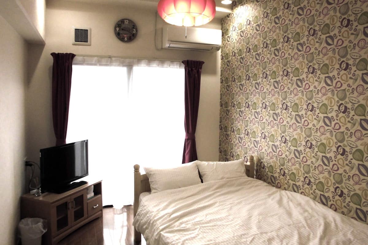 A bedroom with a bed, a television on a cabinet, a window with curtains, an air conditioning unit, and a patterned wallpaper.