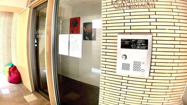 Entrada de un hotel con puerta de vidrio, panel de control digital para el ascensor y una pared de ladrillo con el nombre 'Le Ristorante Zumimachi' grabado.
