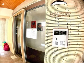 A hotel entrance with a glass door, a digital elevator control panel, and a brick wall with the name 'Le Ristorante Zumimachi' engraved.
