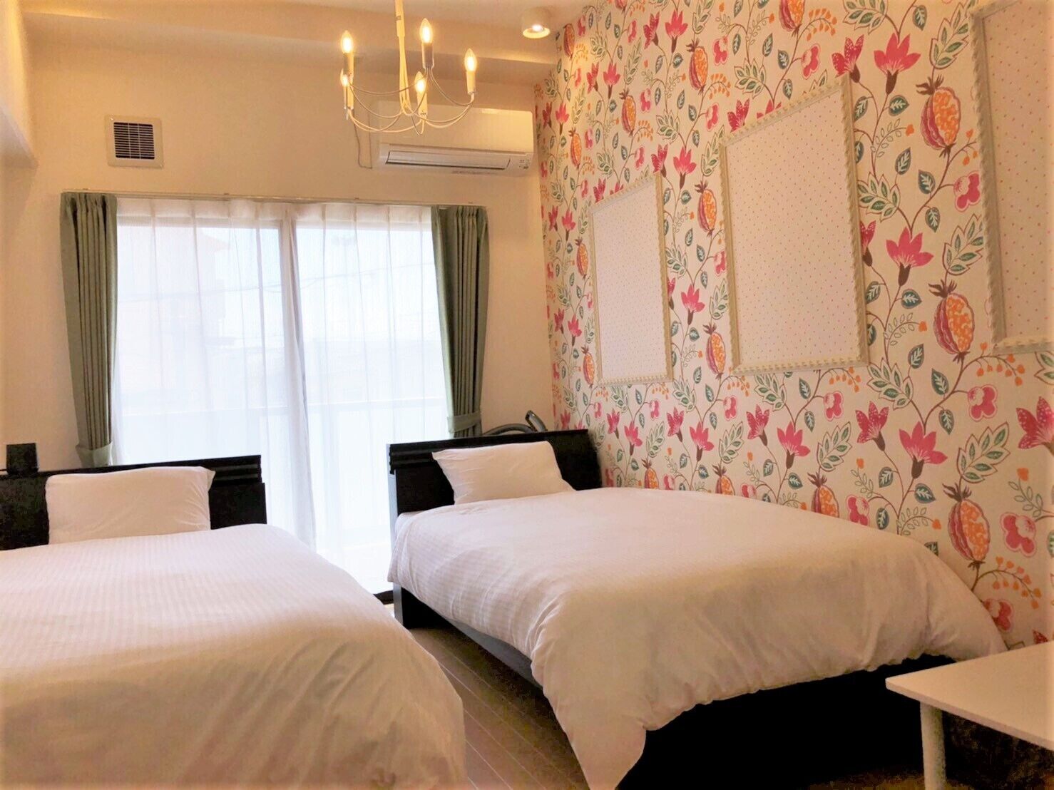 A hotel room with two beds, a chandelier, a window with curtains, and a floral wallpaper design.