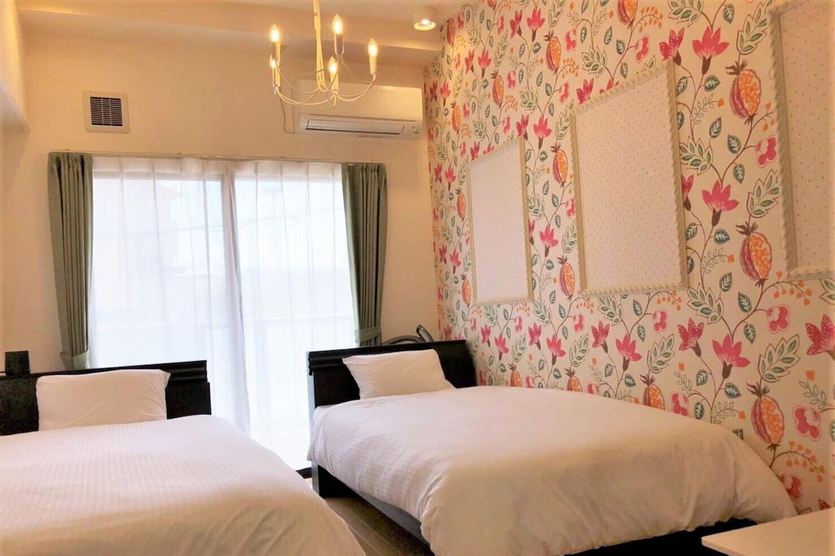 A hotel room with two beds, a chandelier, a window with curtains, and a floral wallpaper design.