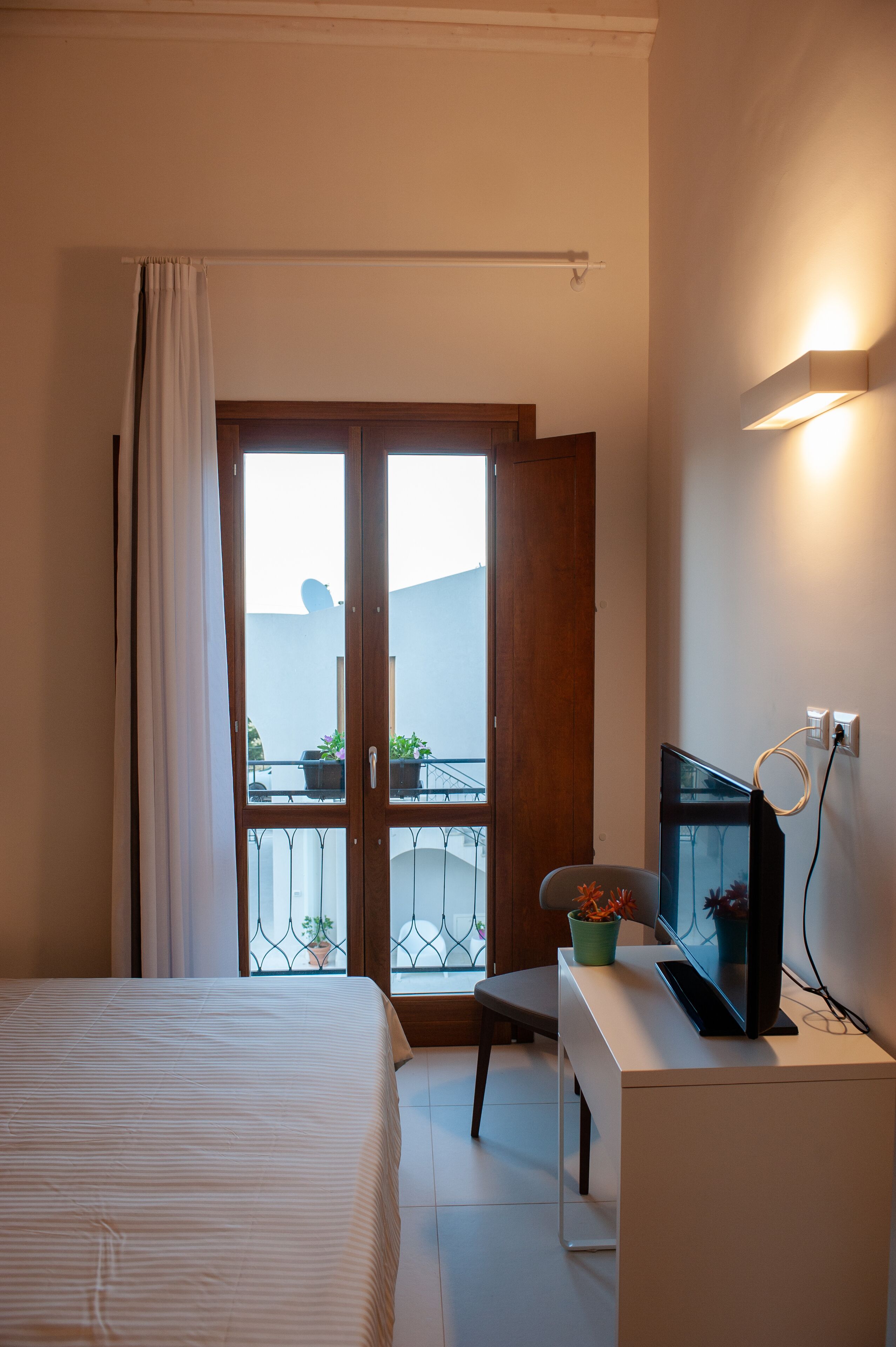 A bedroom with a bed, a desk with a TV, a chair, a balcony with potted plants, and a window with curtains.