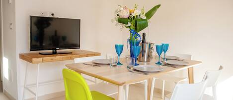A dining area with a wooden table set for four, white chairs, a blue vase with flowers, and a television on a wooden stand.