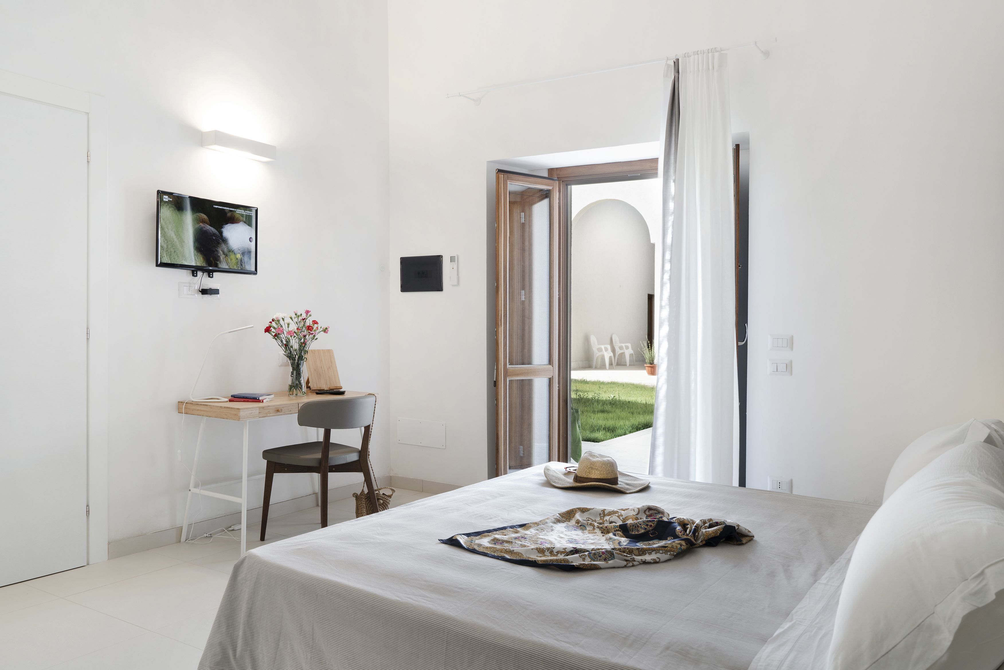 A bedroom with a bed, a desk, a chair, a TV, and a view of a garden.