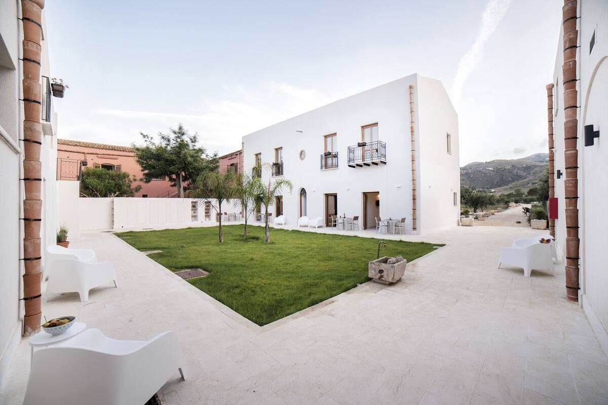 A courtyard with white chairs, a small lawn, and a building with balconies.