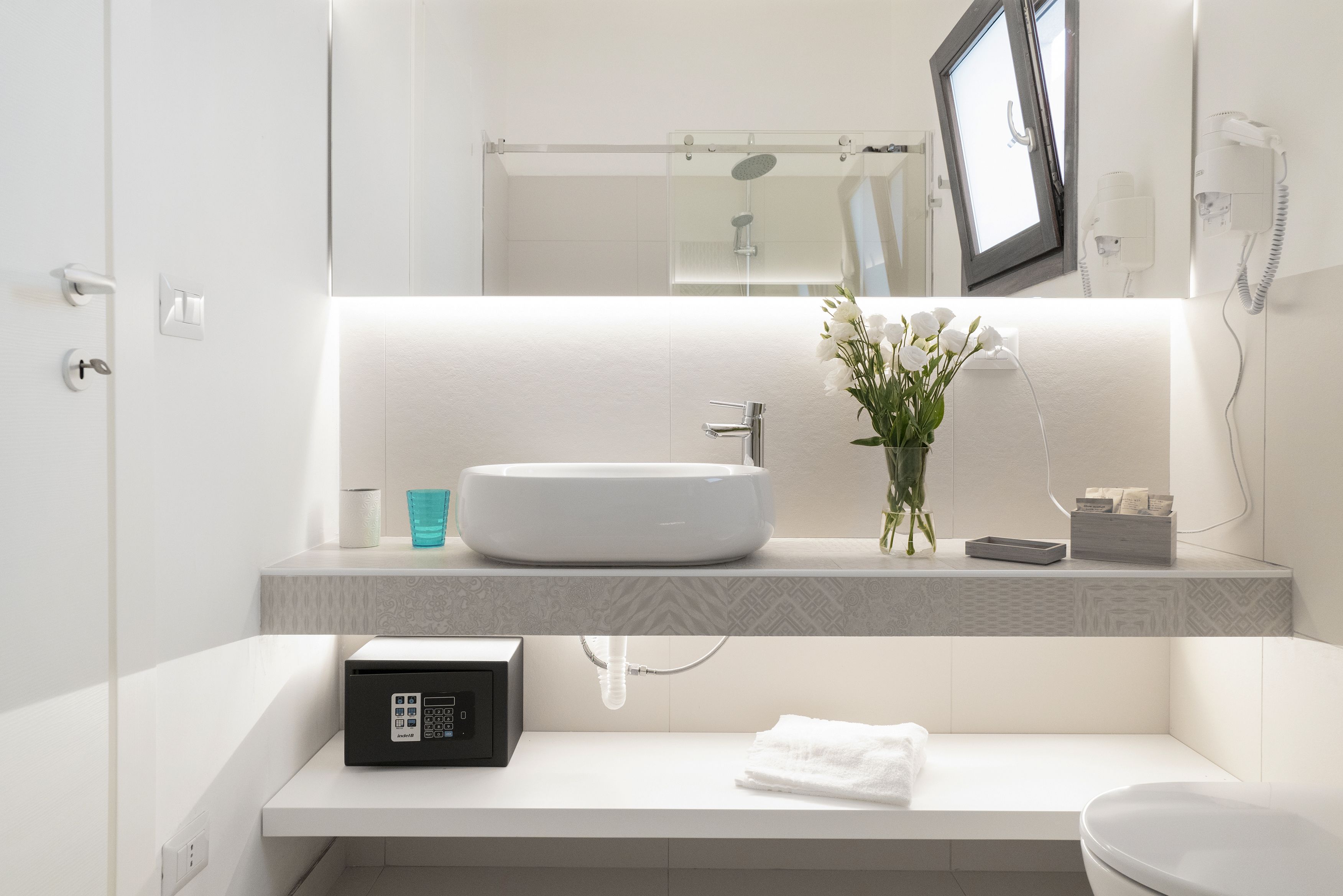A modern bathroom with a white sink, a handheld showerhead, a towel, and a hairdryer on the wall.