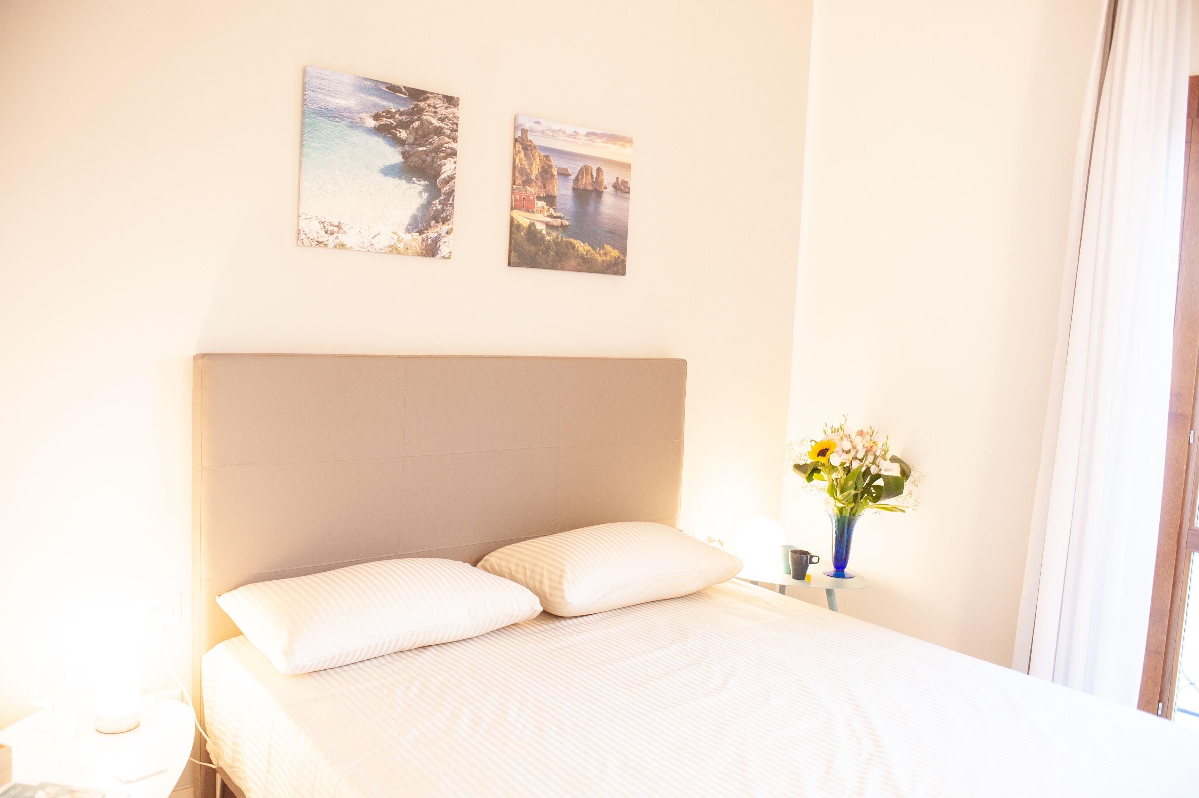 A hotel room with a large bed, two wall-mounted pictures, a vase with flowers, and a window with curtains.
