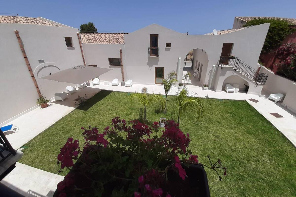 A courtyard with a lawn, white lounge chairs, and a small garden with pink flowers.