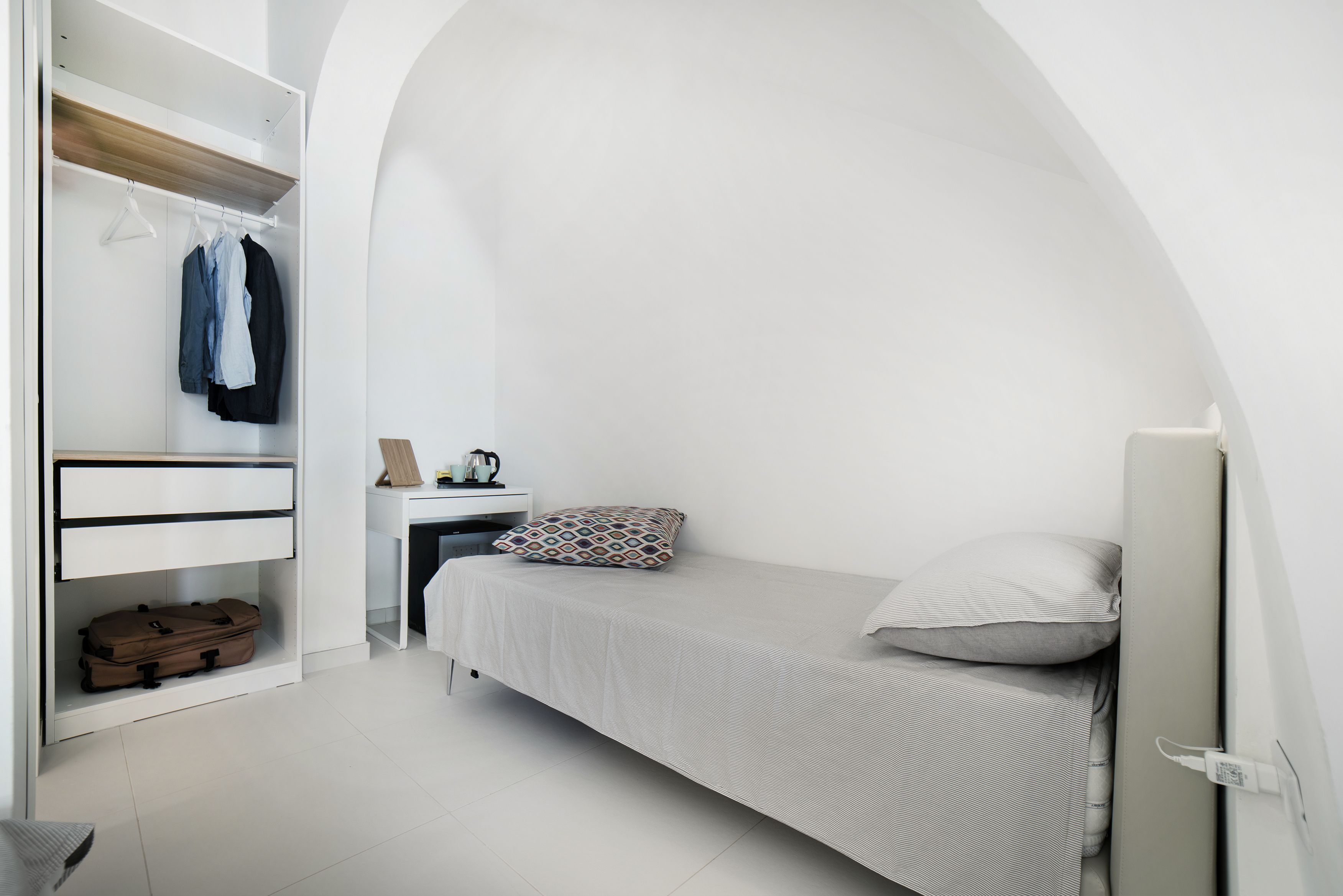 A small, white bedroom with a single bed, a desk, and a wardrobe.