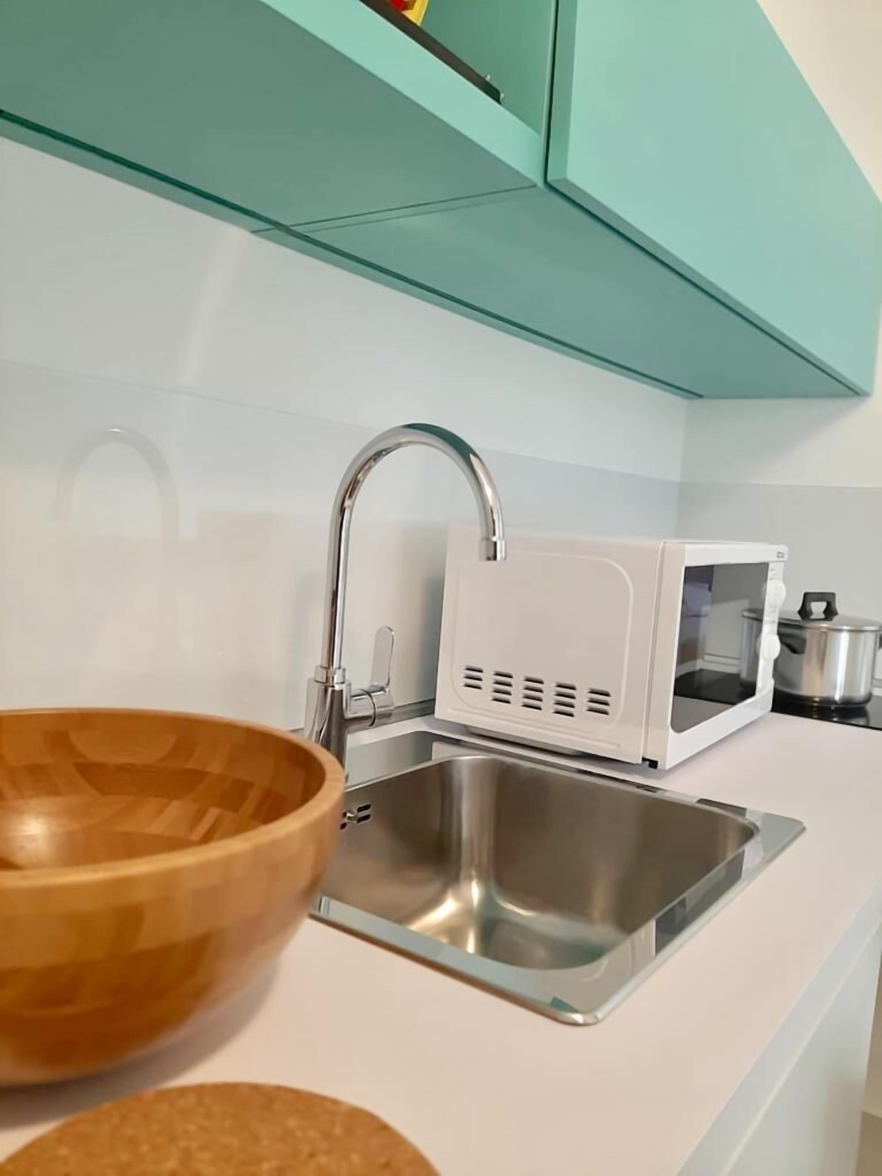 A kitchen with a stainless steel sink, a wooden bowl, a white microwave, and teal cabinets.