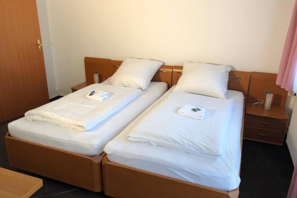 A double bed with white linens and pillows, a wooden headboard, a bedside table with a lamp, and a door in the background.