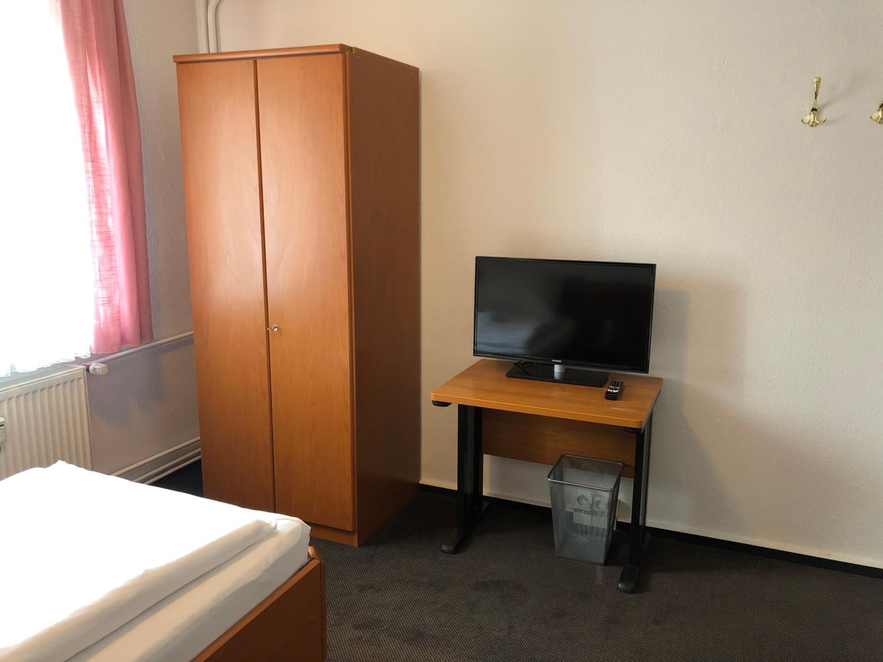 A hotel room with a wooden wardrobe, a bed with white bedding, a television on a wooden stand, and a small trash bin.