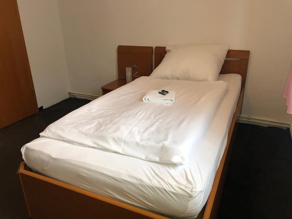Una cama bien hecha con sábanas blancas y una toalla doblada encima, un cabecero de madera y una mesita de noche con una lámpara.