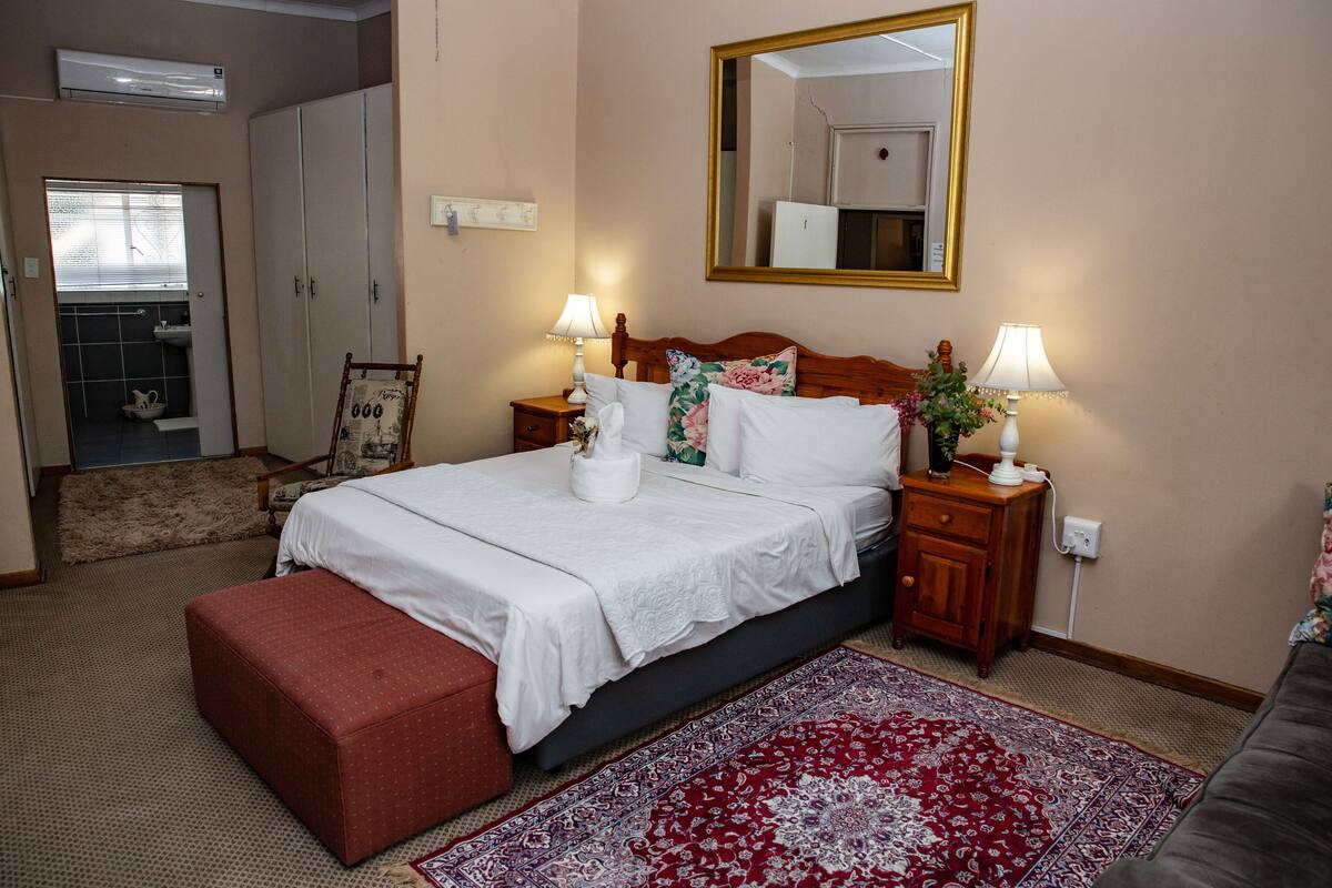 A hotel room with a bed, two bedside tables with lamps, a red ottoman, a large mirror, and a red and white patterned rug.