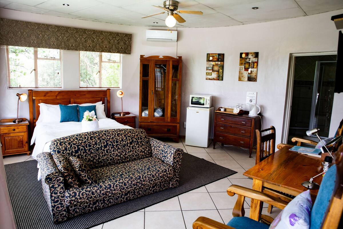 A bedroom with a bed, a wooden wardrobe, a desk, a chair, a microwave, and a ceiling fan.