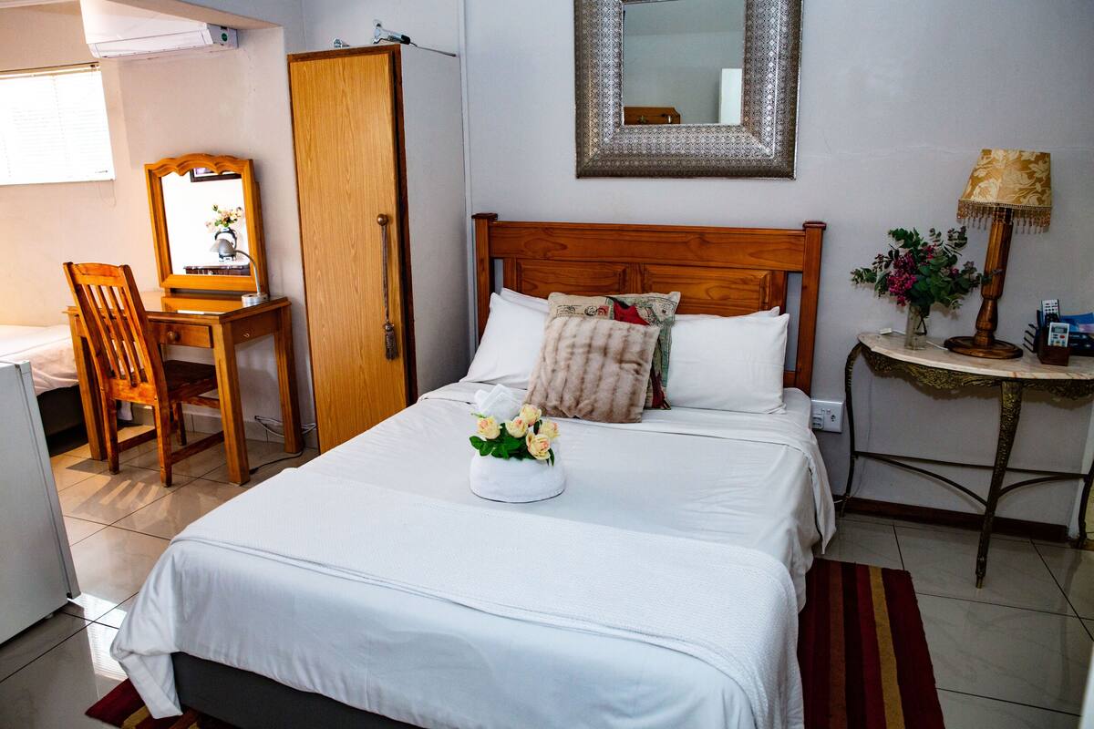 A neatly arranged hotel room with a bed, a wooden chair, a desk, and a small table with a lamp.