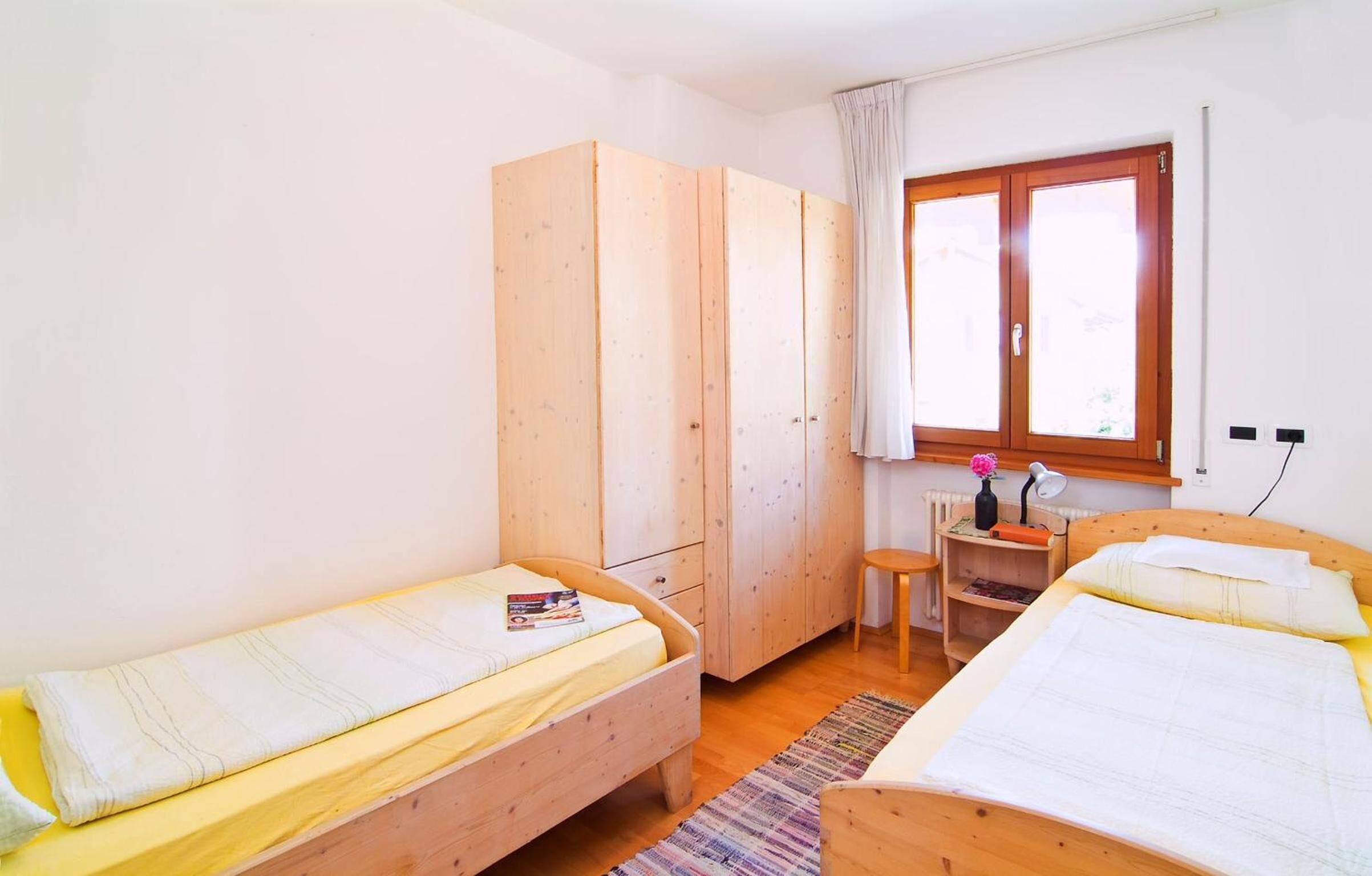 A room with two single beds, wooden wardrobe, desk with chair, and a window with curtains.