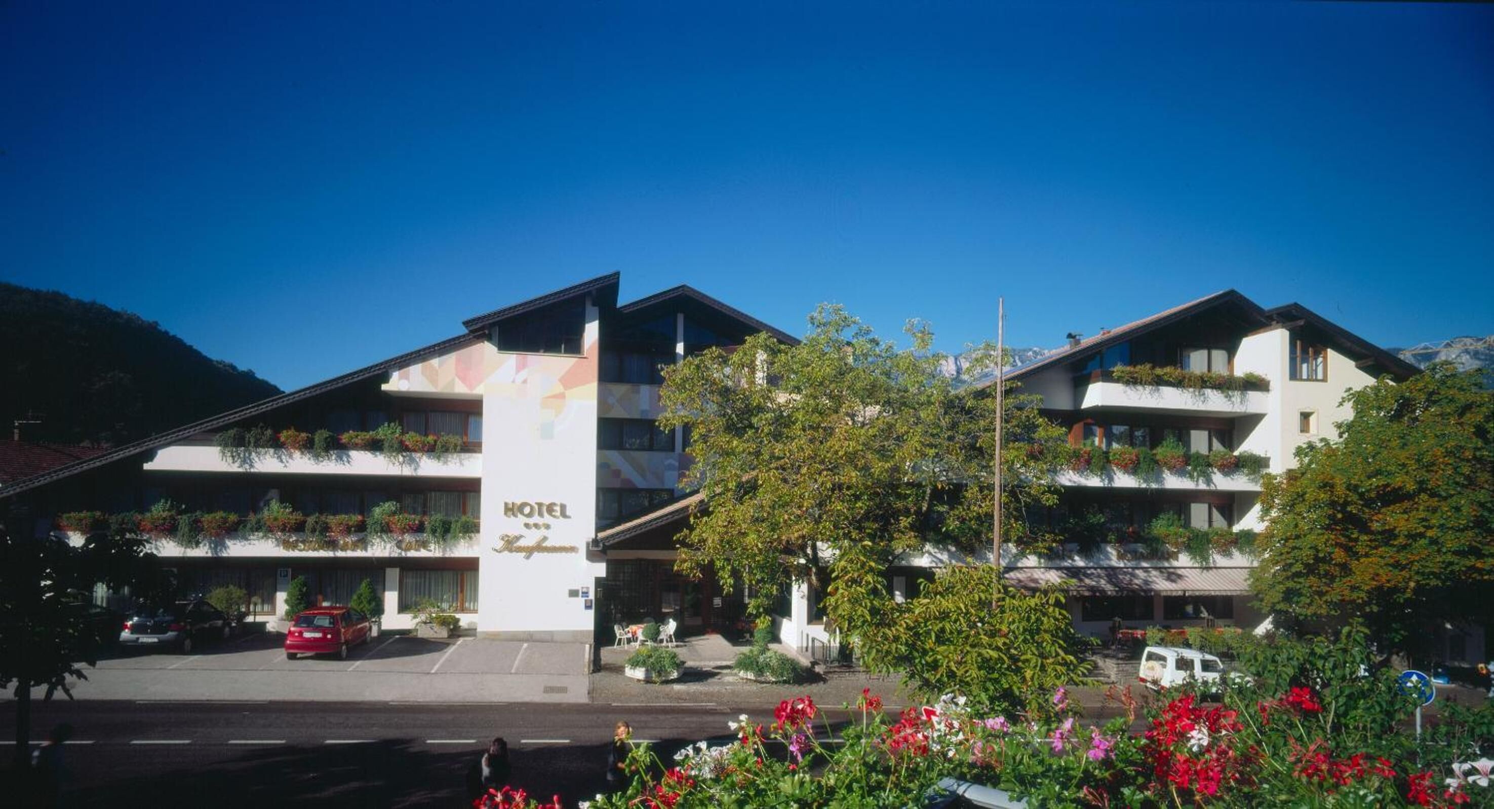 A hotel with a modern design, featuring balconies with plants, a clear sky, and a parking area.