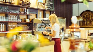 A barista preparing coffee at a counter with a coffee machine, glassware, and a menu board in a cafe setting. - Bio-Hotel & Residence Kaufmann (Ora)