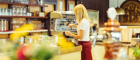 A barista preparing coffee at a counter with a coffee machine, glassware, and a menu board in a cafe setting.