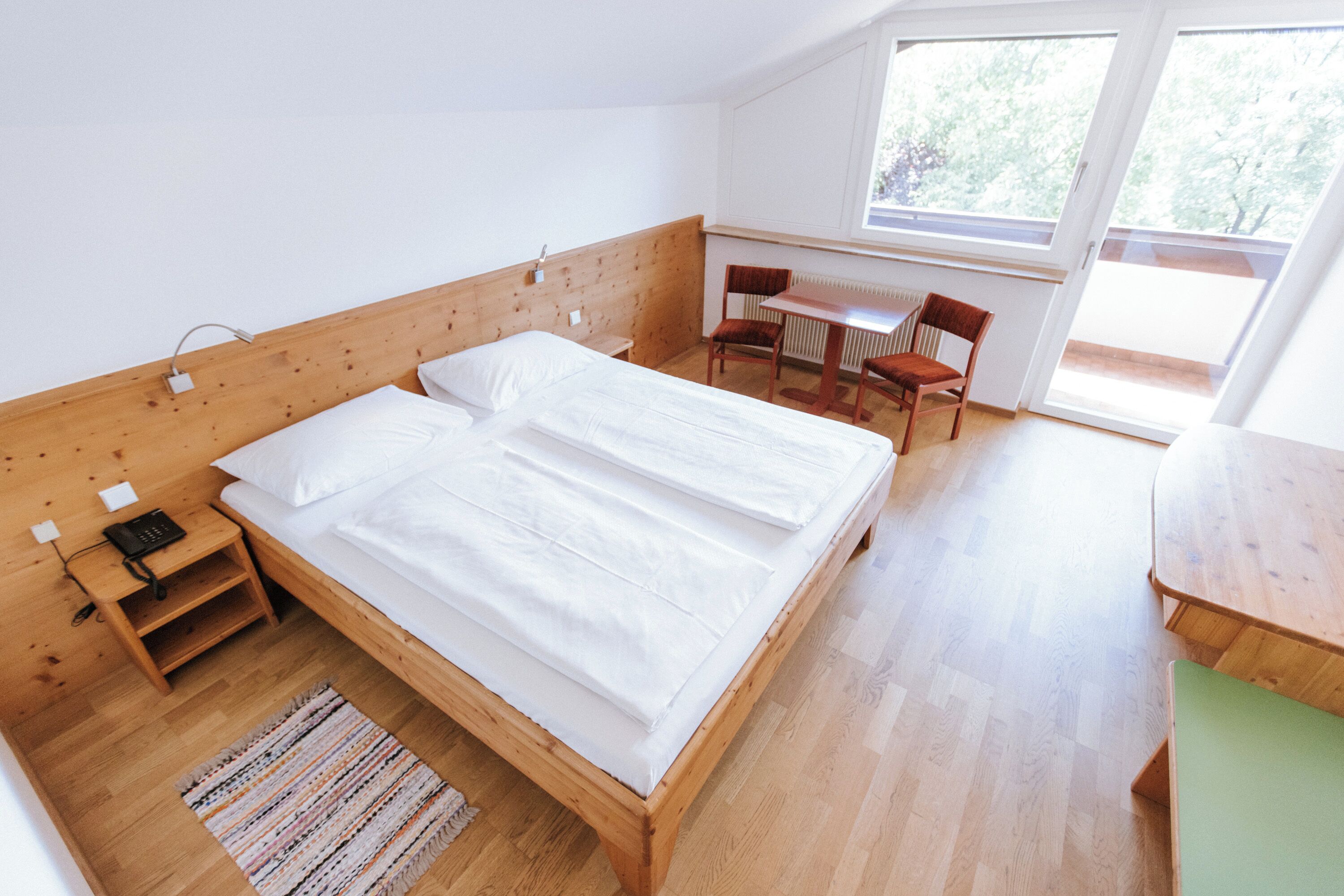 A room with a wooden bed, a small table, a chair, and a window.